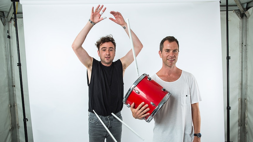 Two men, one wearing a black tee and the other a white tee holding a red drum, stand in front of a white backdrop.