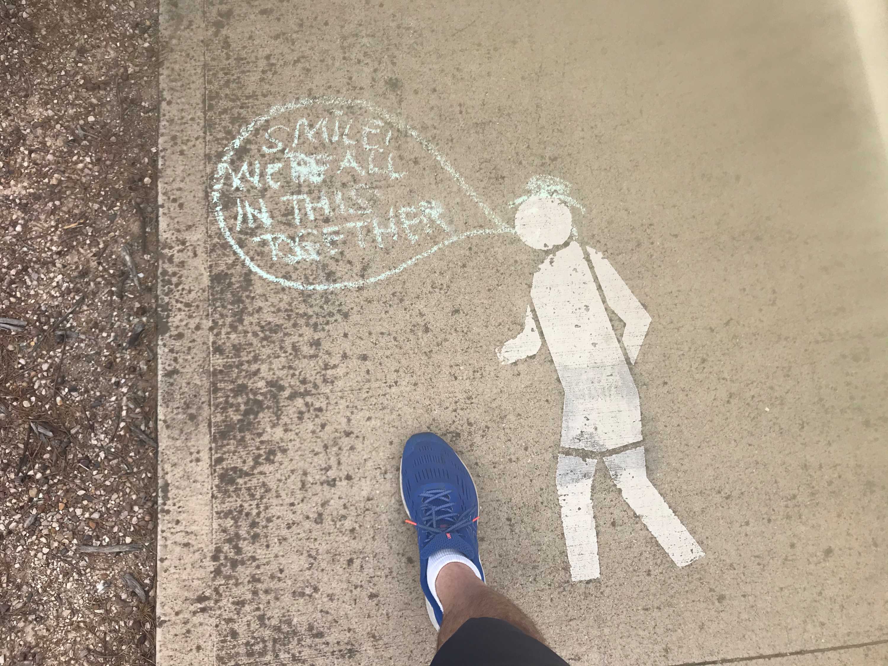 A speech bubble written in chalk on a footpath walking sign saying 'smile, we're all in this together'.
