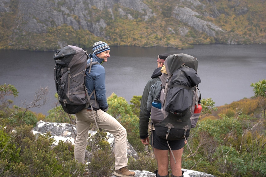 Two men wearing hiking gear, carrying heavy back backs smiling at a lake lookout. 