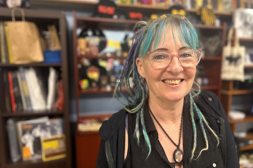 A woman with blue strands in her hair smiles as she stands in her record shop with records in the background