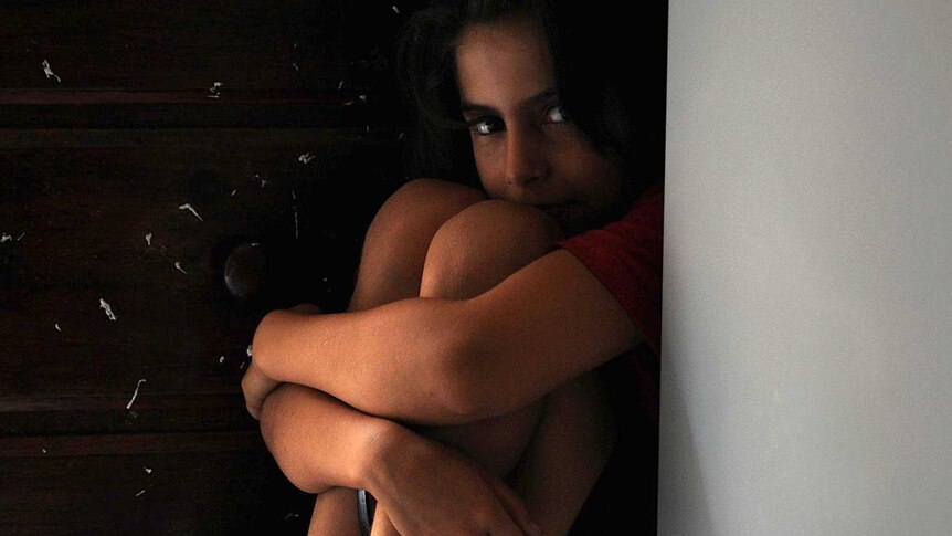 A young child in shadow sits in a corner hugging her knees.