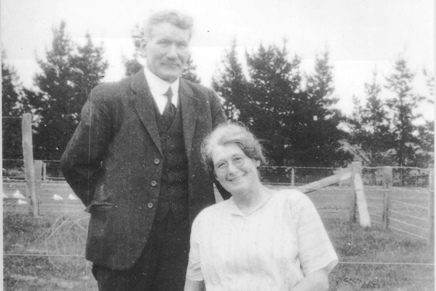 Black and white photo of a Man standing next to his wife sitting in a paddock in front a of wire fence.