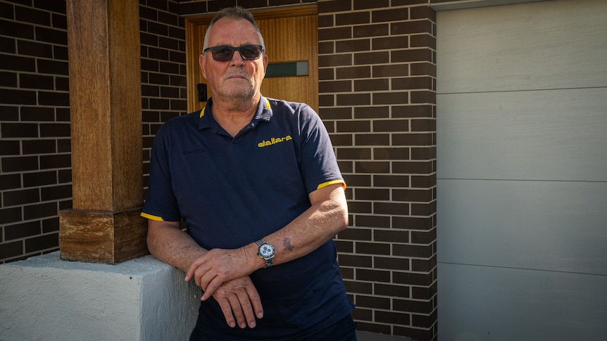An older man wearing a navy blue polo shirt leans against a pillar outside his home wearing sunglasses with his arms folded.