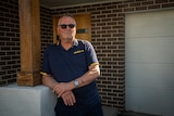 An older man wearing a navy blue polo shirt leans against a pillar outside his home wearing sunglasses with his arms folded.