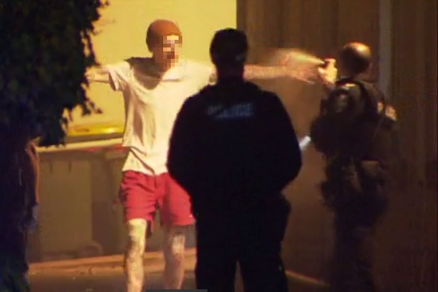 Two police officers spray at a man wearing a white shirt, red shorts and a beanie