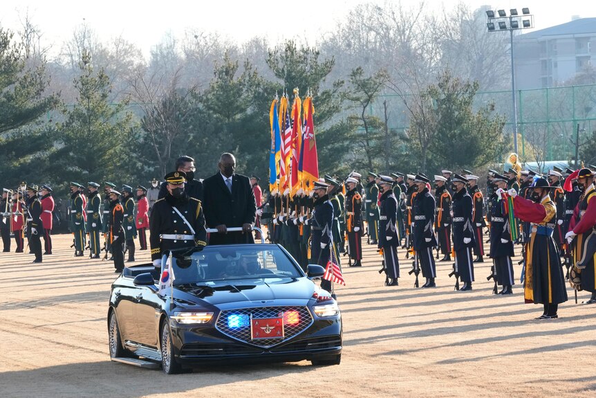 Two men and a guard stand in a ride on car as it drives past military officers performing a guard of honour on a misty day 