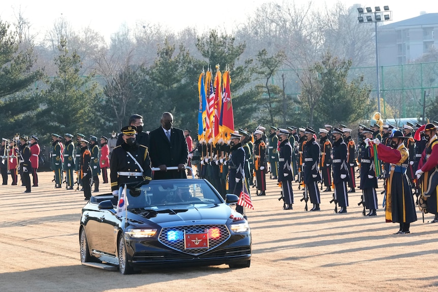 Two men and a guard stand in a ride on car as it drives past military officers performing a guard of honour on a misty day 