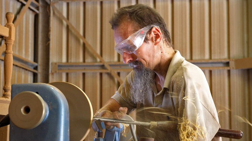 Dale Cordwell holds a wood turning implement while using a lathe, sawdust chips are flying out behind him.
