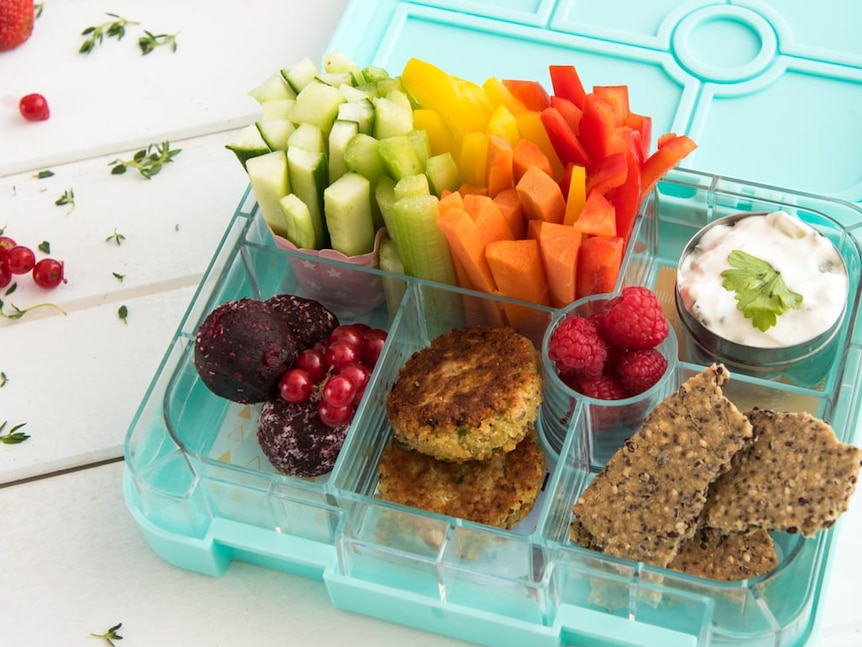 A green lunch box filled with chopped up vegetables, yoghurt, berries, homemade crackers, fritters and energy balls.
