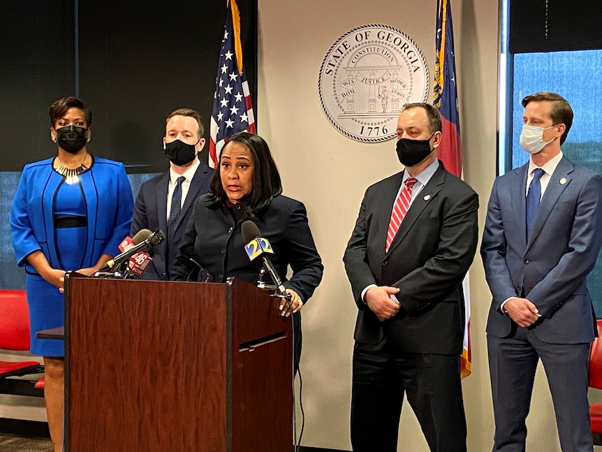 A black woman in a black suit stands  behind a wooden podium with a silver sign behind that says "State of Georgia".