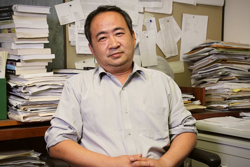 A man sits in a chair in an office surrounded by books and papers