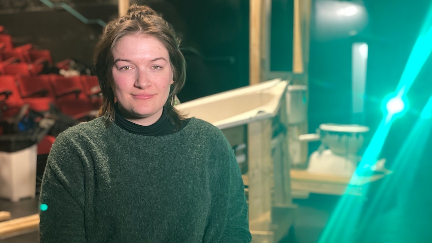 A woman smiles at the camera with theatre props and lighting int he background.