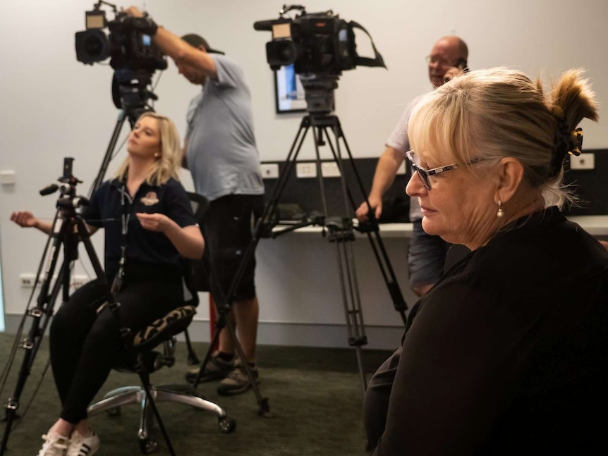 A female Auslan interpreter sits while waiting for a press conference to begin. Cameras are being set up in the background.