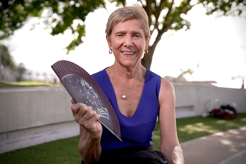 A woman in a blue shirt fans herself with a handheld fan as she smiles at the camera.