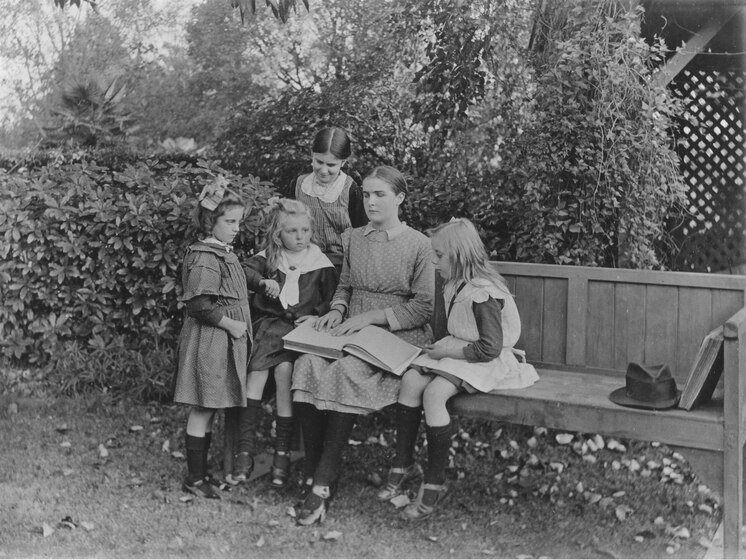 A black and white photograph showing a woman sitting on a bench with a book open, with four children around her.