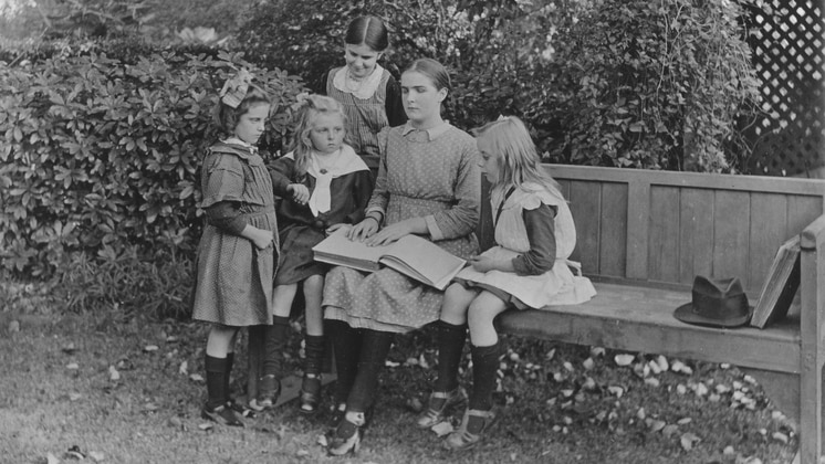 A black and white photograph showing a woman sitting on a bench with a book open, with four children around her.