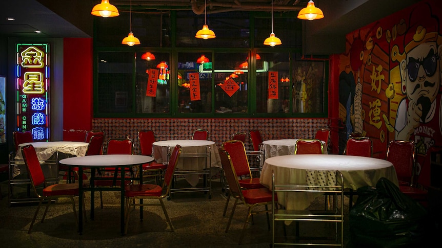 Empty red seats placed at tables covered in white table cloths below yellow lamps and neon signs