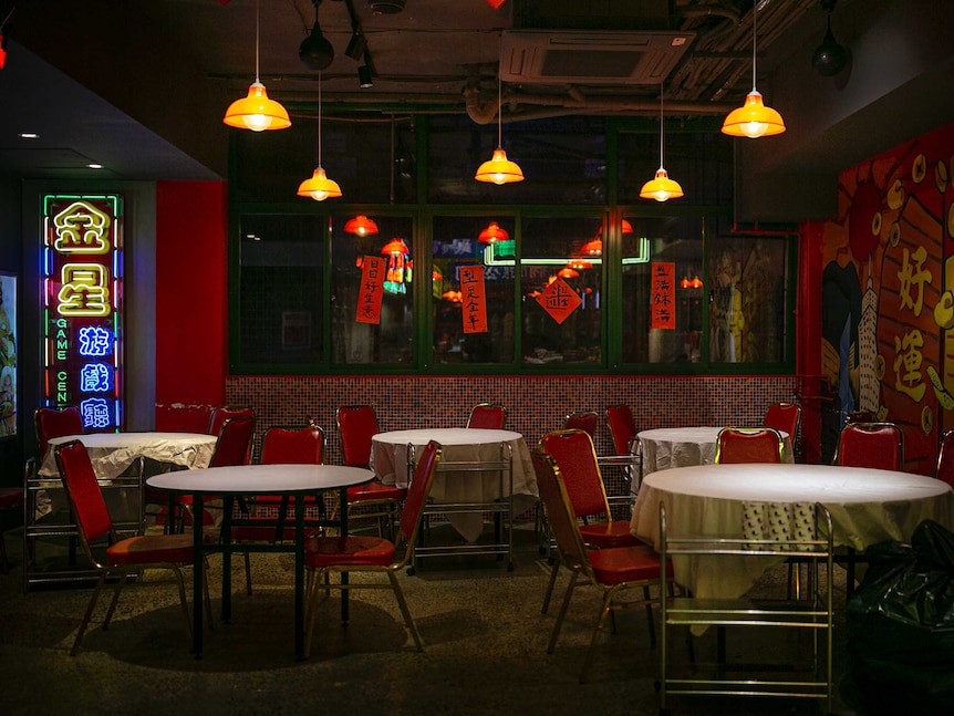 Empty red seats placed at tables covered in white table cloths below yellow lamps and neon signs