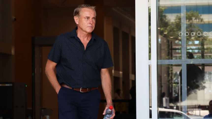 A man wearing a dark blue shirt and pants walks out a sliding door with his hand in his pocket