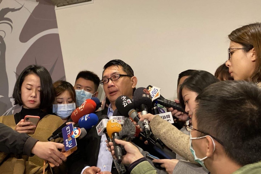 Vincent Hsu speaks to reporters who are holding several microphones near his face.