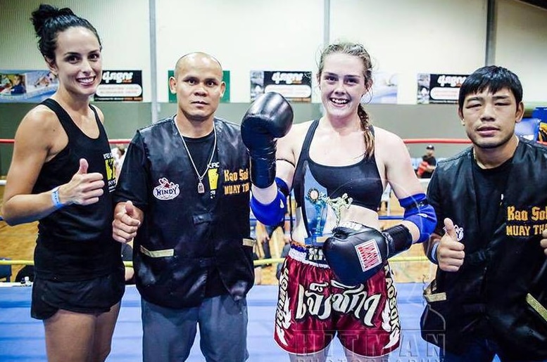 A woman wearing kickboxing gear stands in a ring with other people flashing a thumb's up.