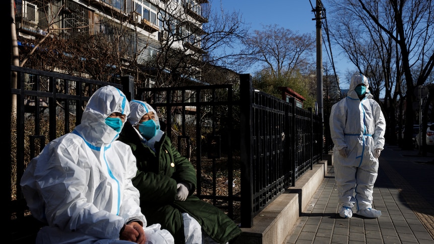 three people in white protective suits stand outside a building's black fence in daytime
