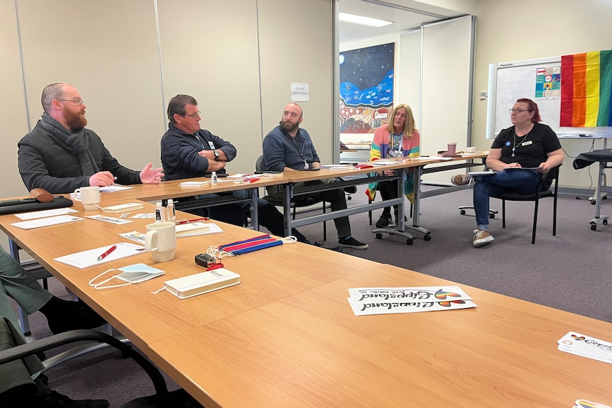 Table in foreground with Gippsland Pride flag in background, four participants sit talking in an office.