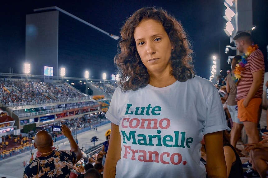 A woman wearing a shirt with a message printed on it stands among a large crowd of spectators in stadium seating