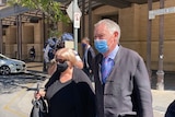A woman with short blonde hair in a black top and mask walks beside a man with grey hair in a suit and a mask outside court