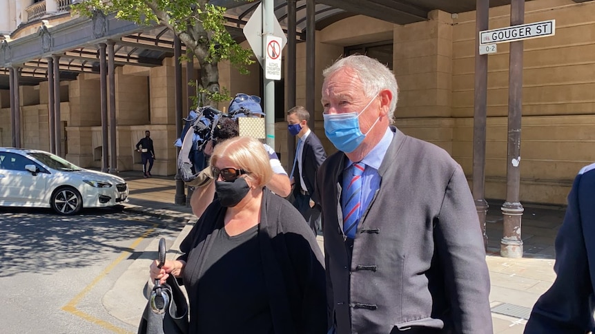 A woman with short blonde hair in a black top and mask walks beside a man with grey hair in a suit and a mask outside court