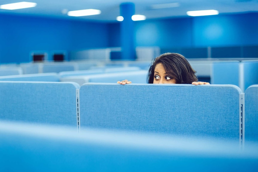 A woman ducks behind a blue cubicle. Only her wide eyes and dark brown hair can be seen over the top of it.