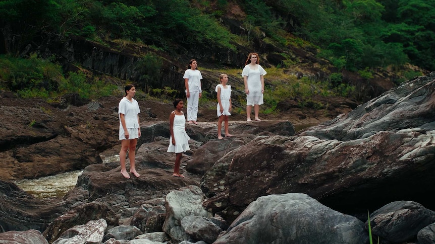 Five young choristers dressed in white sing on rocks along the coastline of Djabugay Country.