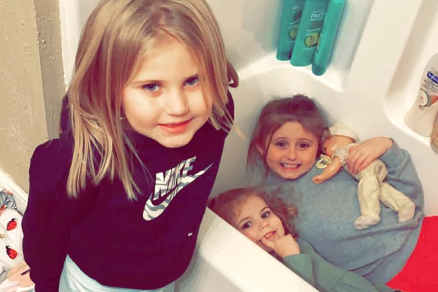 Trois jeunes filles blondes sourient dans leur salle de bain.  Deux sont dans la baignoire, dont l'un tient une poupée.