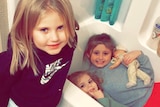 Three young blonde girls smile in their bathroom. Two are in the tub, one of whom is holding a baby doll.