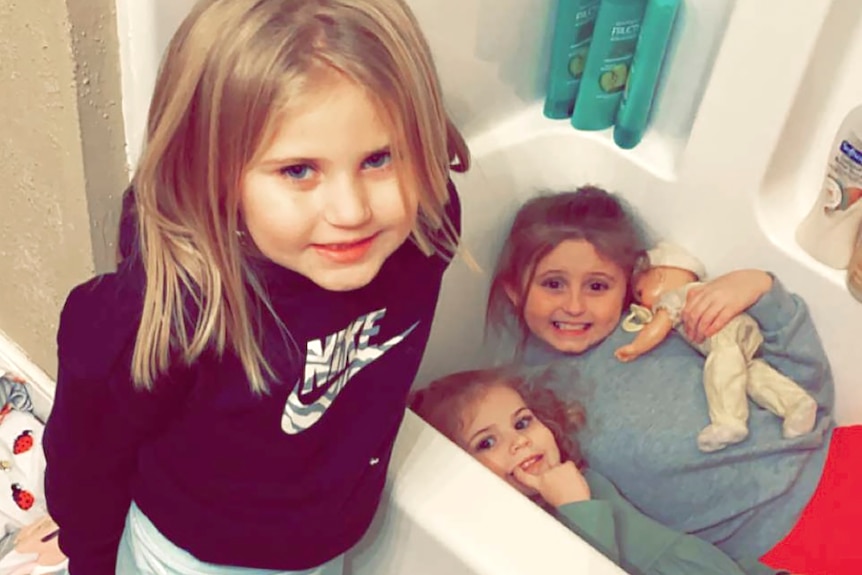 Three young blonde girls smile in their bathroom. Two are in the tub, one of whom is holding a baby doll.