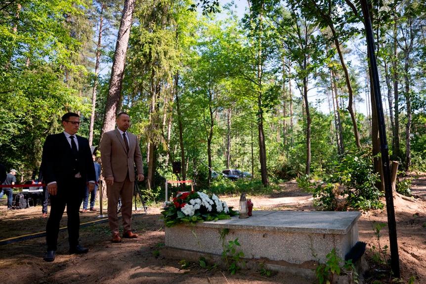 Dwóch mężczyzn w garniturach stoi przed grobem z bukietami kwiatów w środku lasu. 