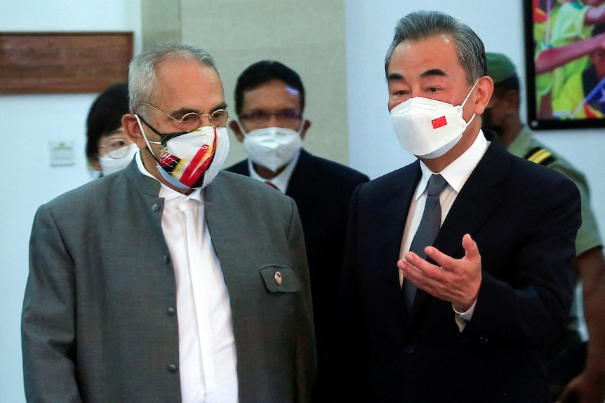 Two men in suits and face masks talk inside a building.