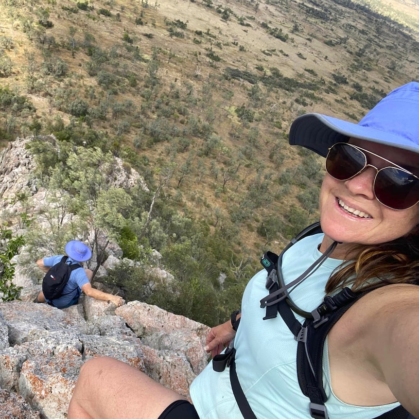 A woman smiles taking a selfie on a mountain with a man climbing below