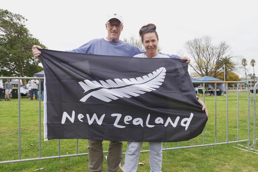A man and a woman standing behind a black New Zealand flag with the silver fern 