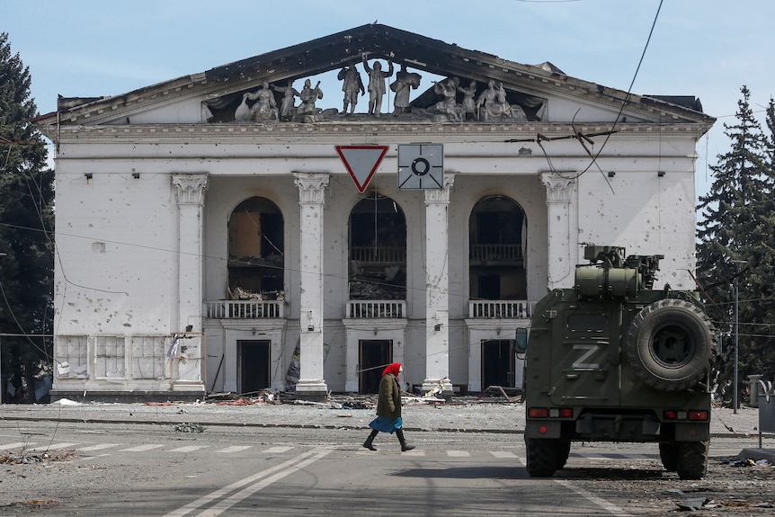 A woman walks next to an armoured vehicle and a destroyed theatre building