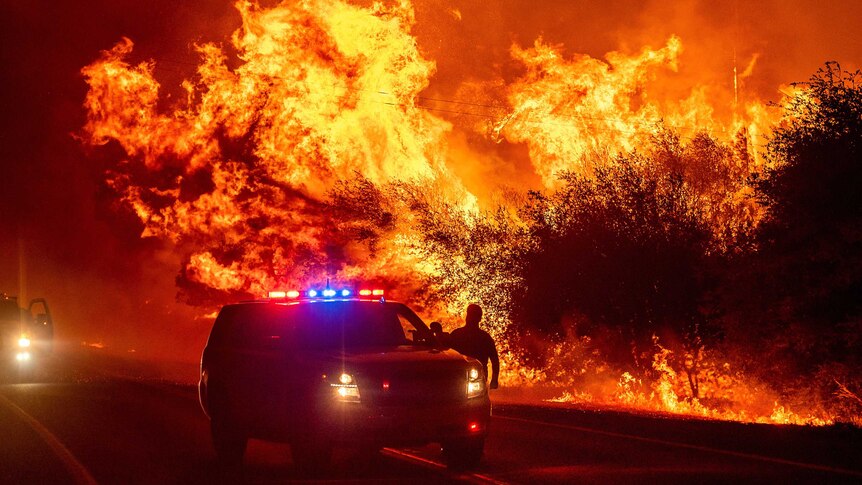 Flames burn bright orange right behind an emergency vehicle as a man stands by the car door.