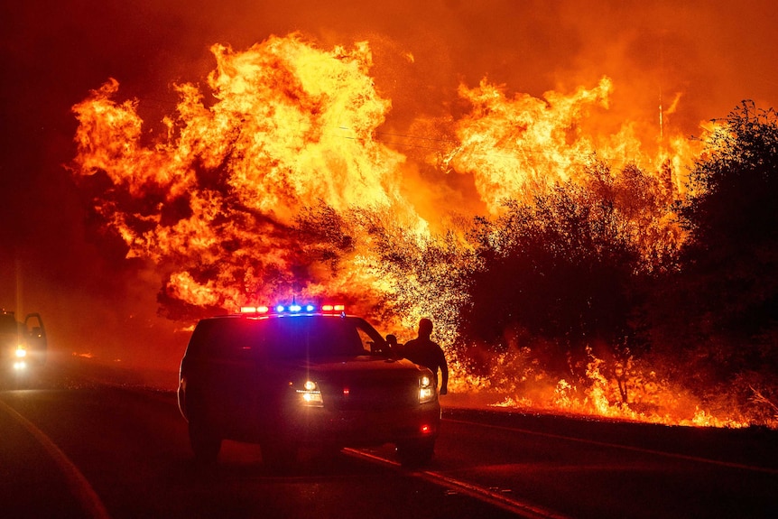 Flames burn bright orange right behind an emergency vehicle as a man stands by the car door.