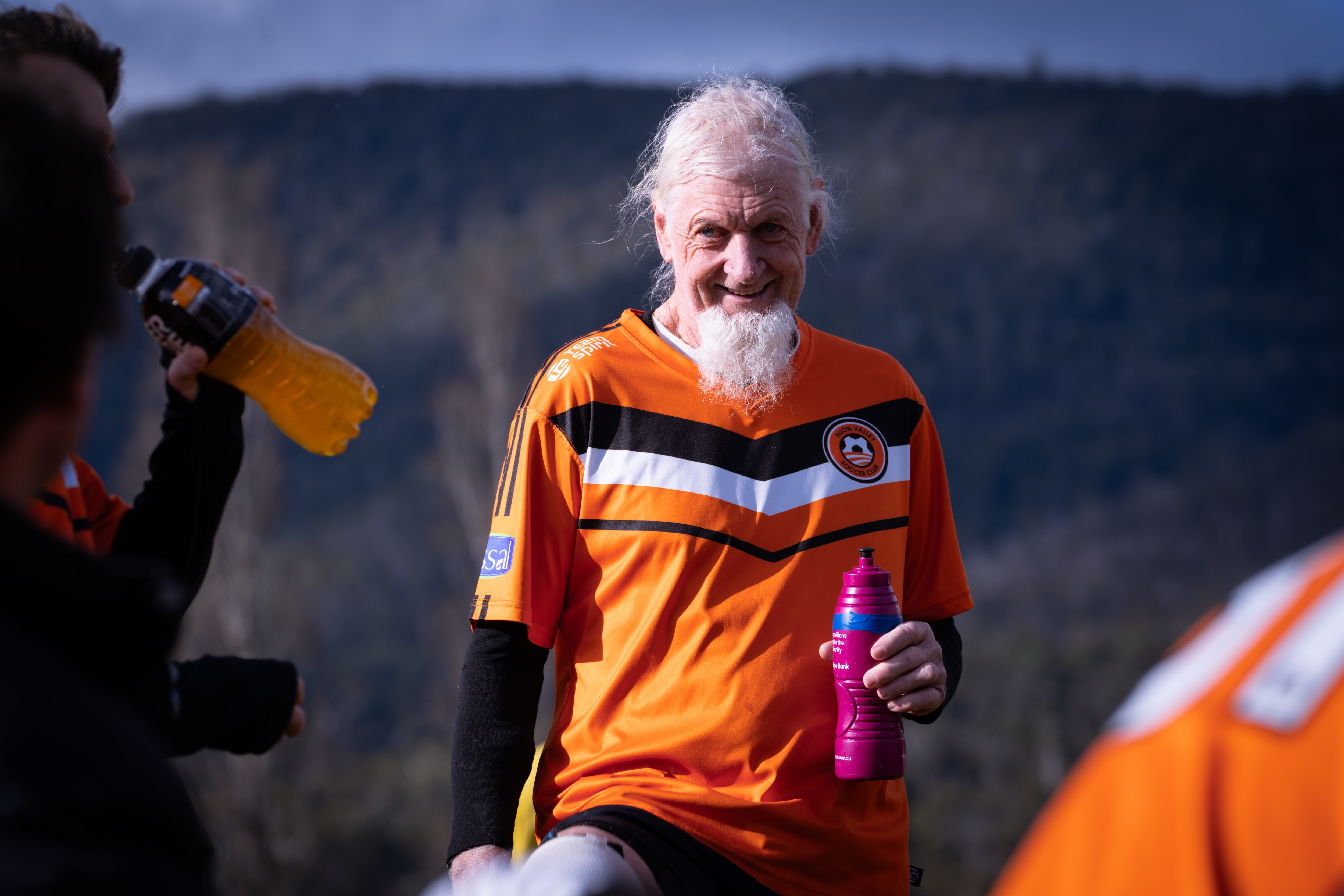 A man with white hair and a long white beard wearing orange soccer colours on a soccer field.