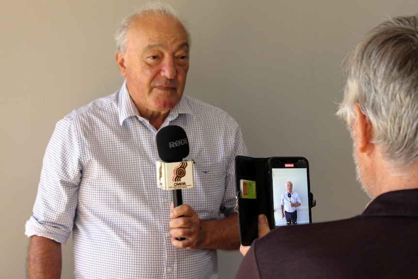 Dr Mike Freelander stands up against a wall holding a microphone as he gets his picture taken