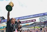 F1 driver Lewis Hamilton shouts in celebration as he hoists a large gold trophy in the air, while a big group of fans watch.