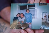 Hands hold a photograph of an older gentleman surrounded by family at a kitchen table.