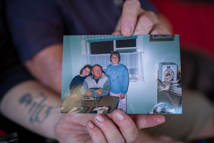 Hands hold a photograph of an older gentleman surrounded by family at a kitchen table.