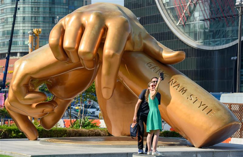 Una pareja de turistas se toma un selfie frente a una enorme estatua de bronce con las manos cruzadas a la altura de las muñecas.