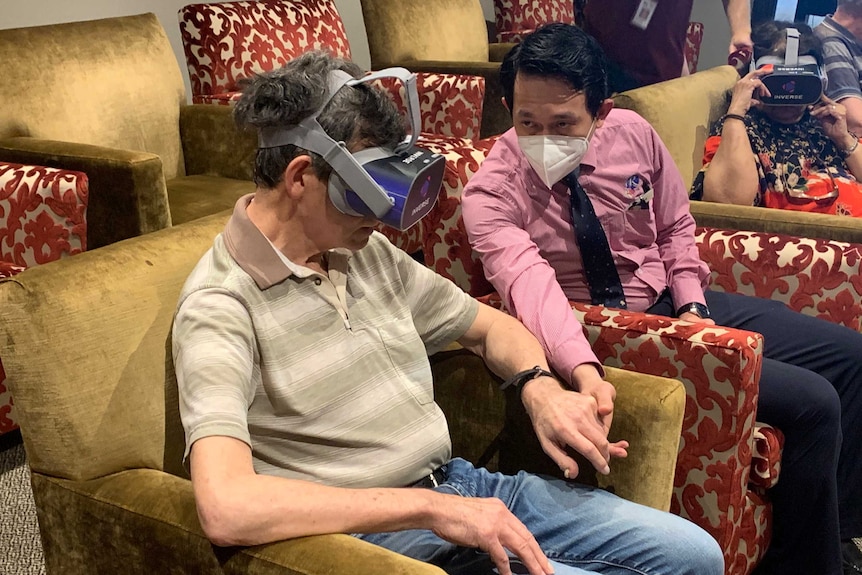 A man in VR goggles sitting down with another man in a surgical mask holding his hand.