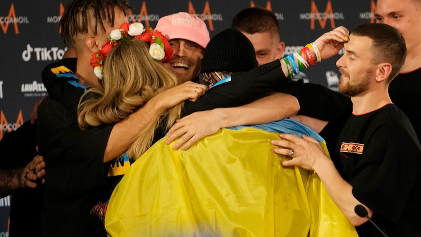 A group of people hug, one man has a Ukrainian flag draped around his shoulders.